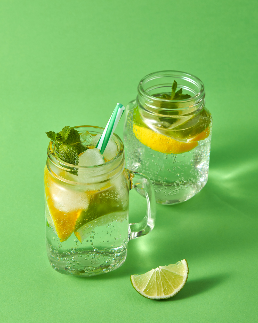 带有气泡的冷天然手工柠檬水两个玻璃梅森罐与绿色背景上的非酒精饮料冷酒精或非酒精夏季饮料的概念夏季清爽的鸡尾酒来自天然柑橘类水果在绿色背景的玻璃杯中加入冰水薄荷和塑料吸管